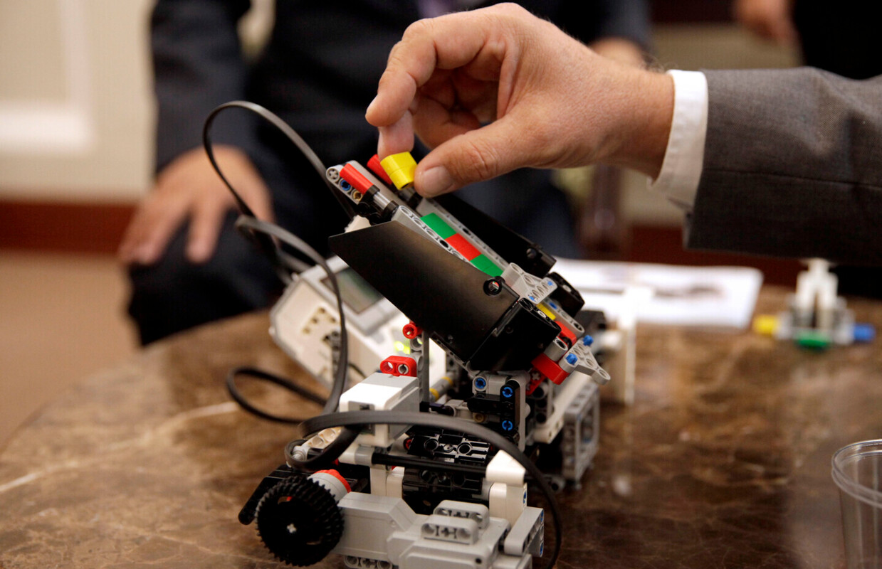 Uma mão a colocar uma peça num protótipo de um robot, pousado numa mesa, atrás vê-se a parte de baixo do tronco de uma pessoa, com uma mão na perna.